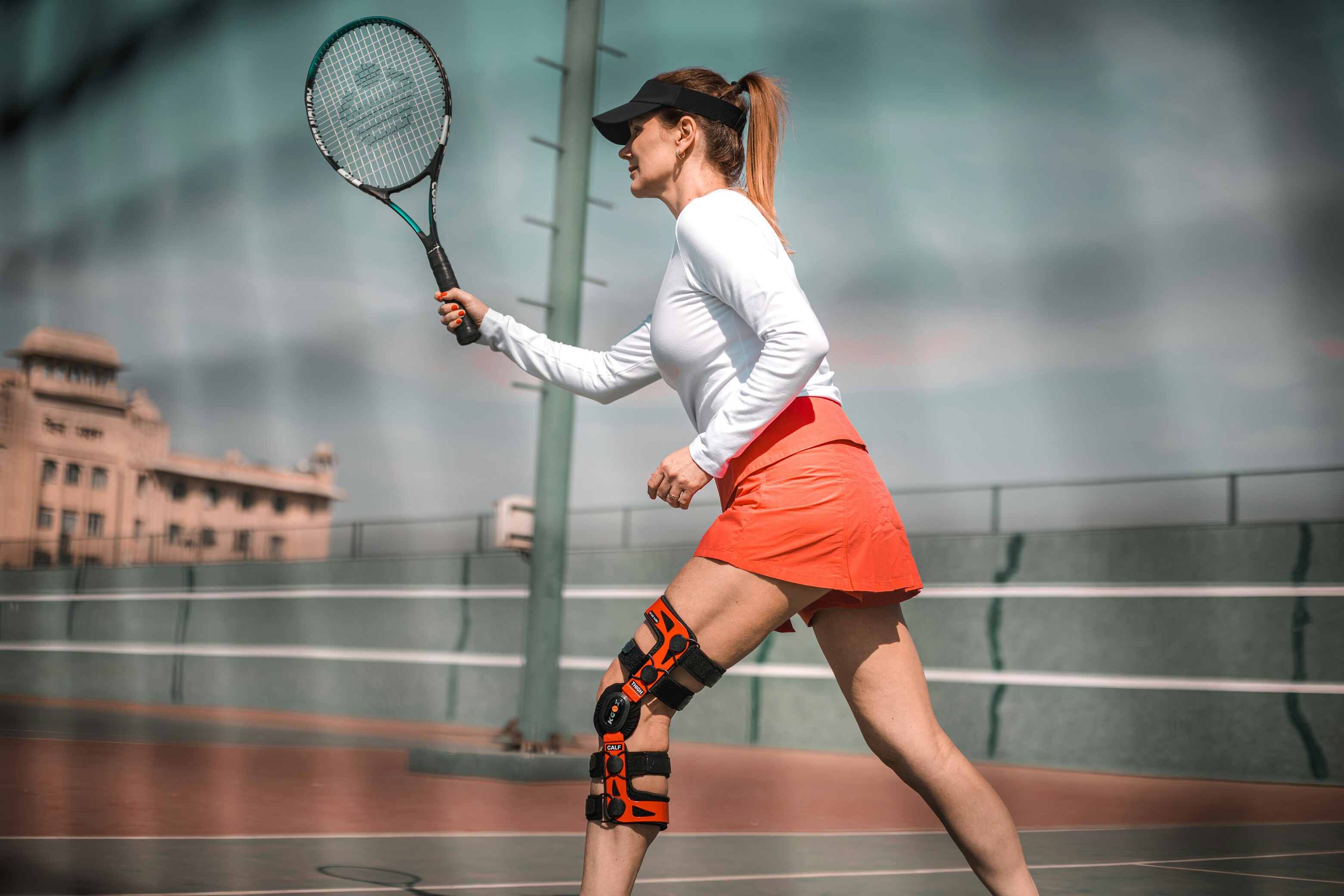 woman in a white top and orange skirt playing tennis on an outdoor court wearing a knee brace