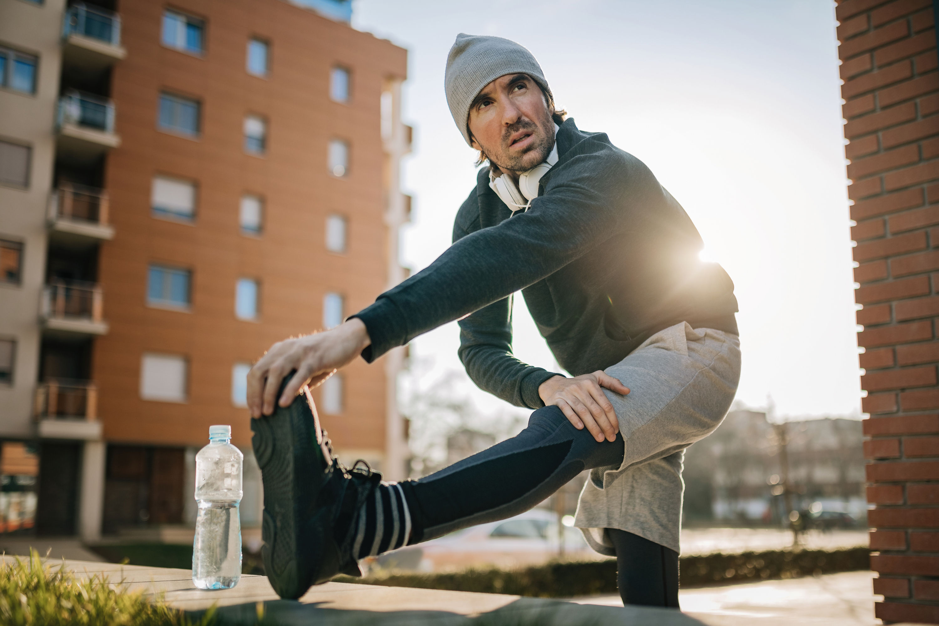 man in a beanie stretching his leg on a bench outside an apartment building with a water bottle nearby